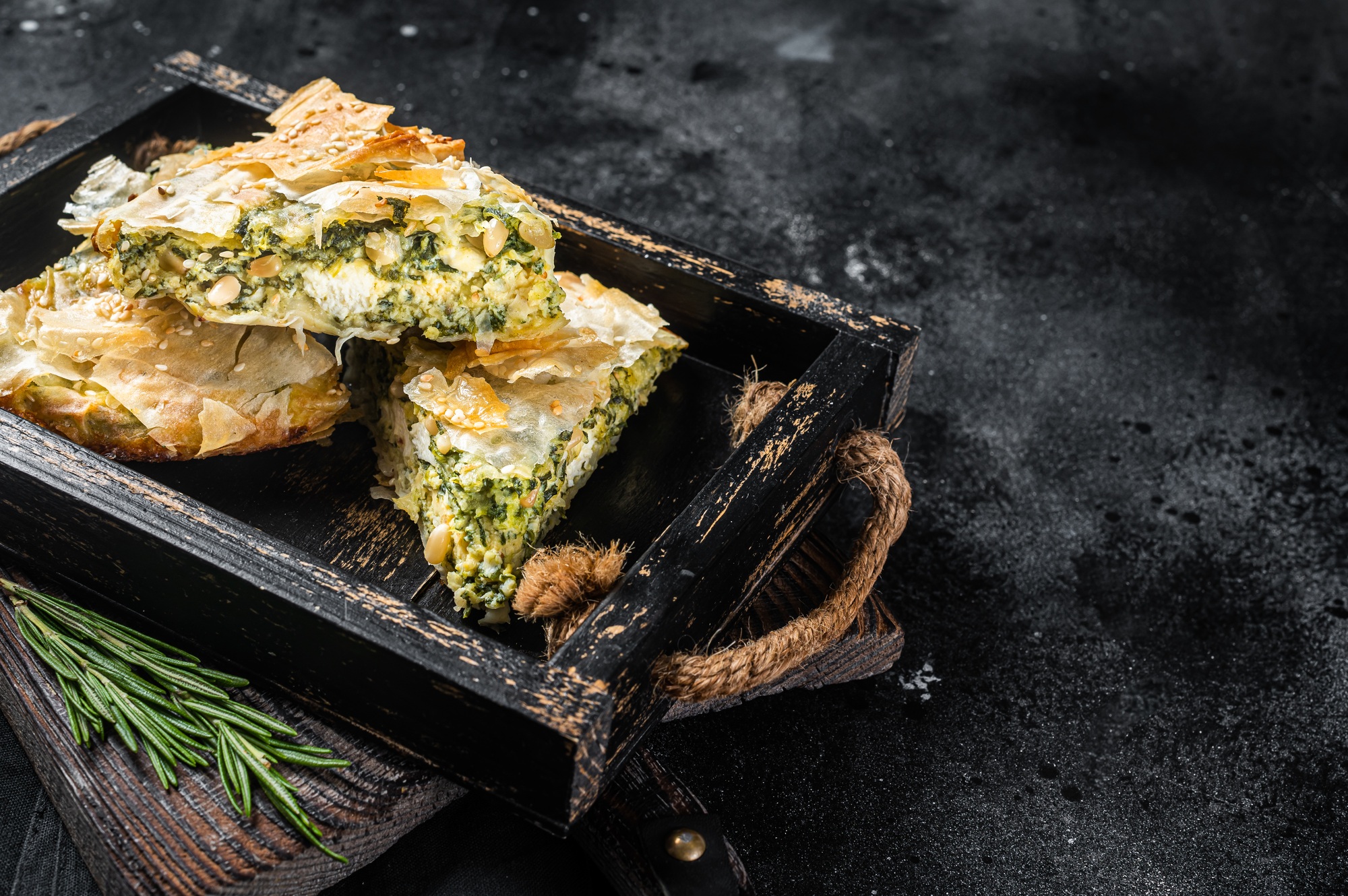 Spinach pie pieces, Spanakopita Greek Pie in wooden tray. Black background. Top view. Copy space